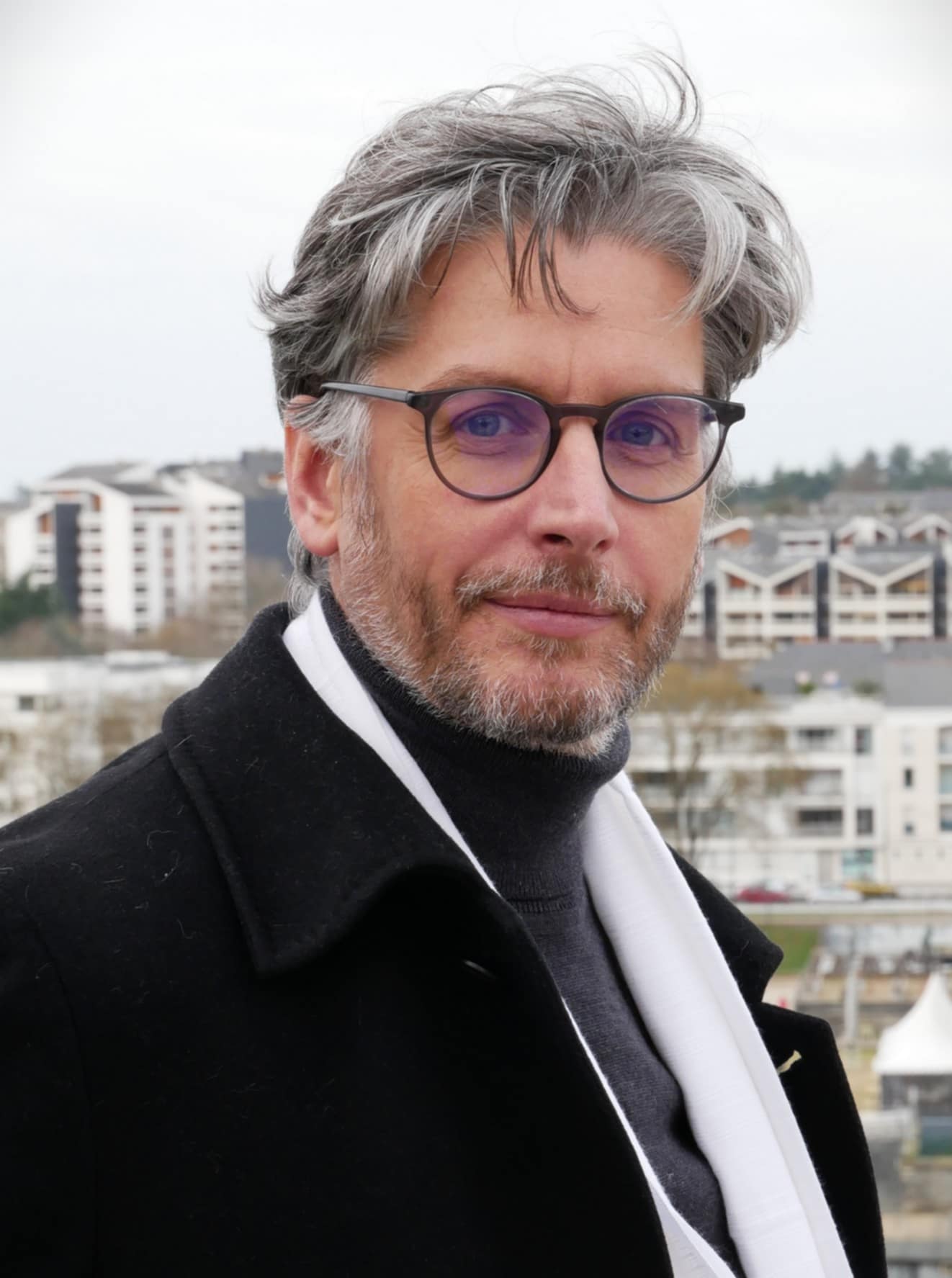 Portrait David Cayla : cheveux gris et aux lunettes, portant un manteau noir et un châle, avec un arrière-plan urbain flou.