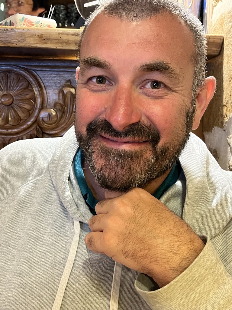 Un homme souriant avec une barbe et des cheveux courts, portant un sweat à capuche gris, s'appuie sur sa main dans un cadre intime.